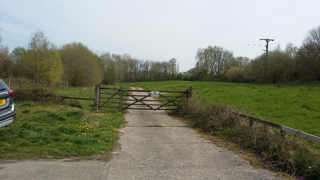 A concrete driveway with grass either side heads off into the distance. There is a wooden gate across the road with a conservation area sign.