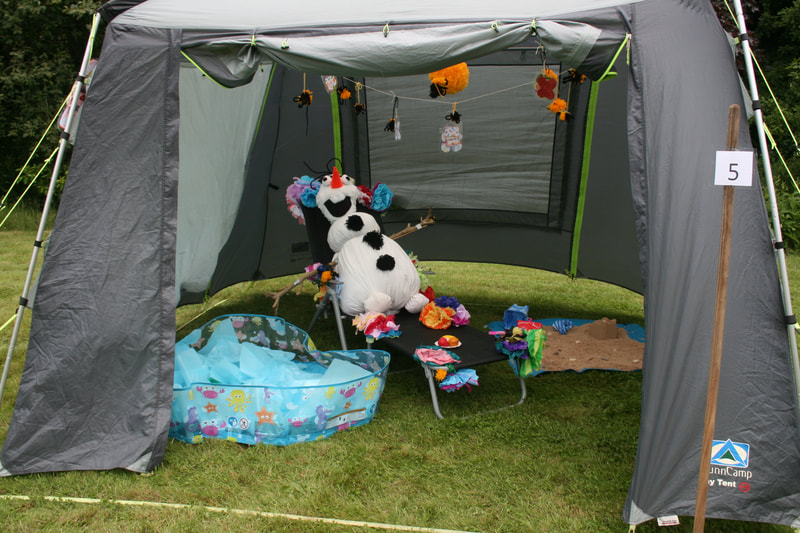 An Olaf scarcrow lounging on a deckchair next to a paddling pool and pile of sand. There is a grey tent sheltering him.